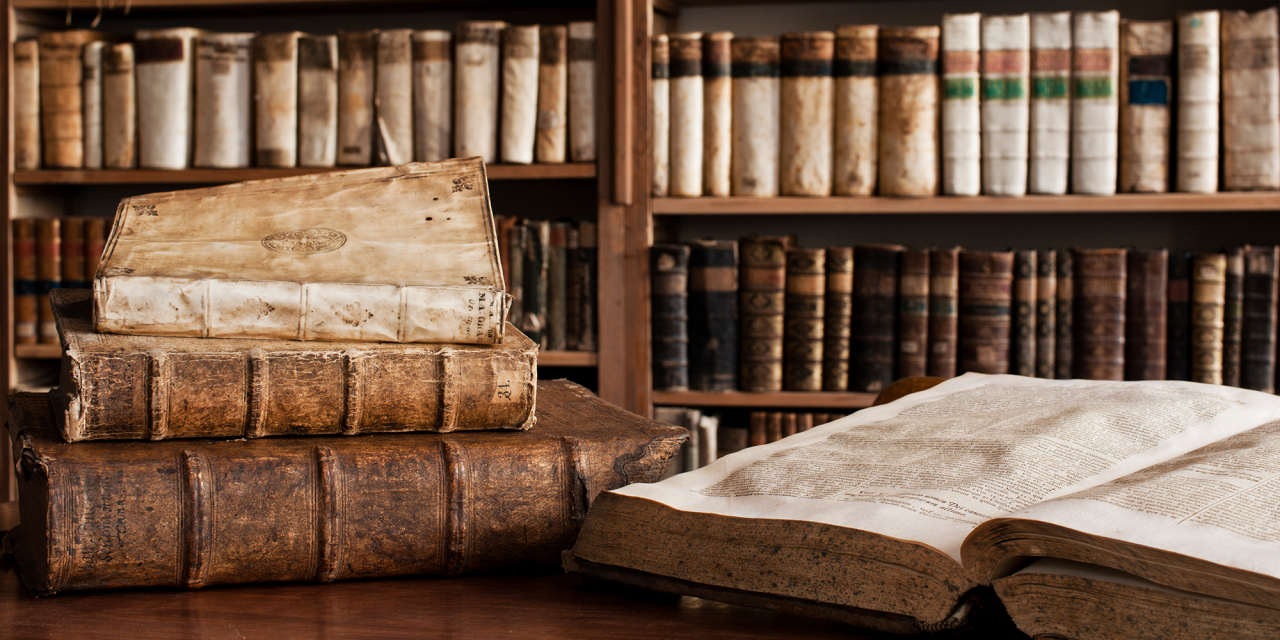 Old books on bookcase and open book next to a stack of books on a desk.