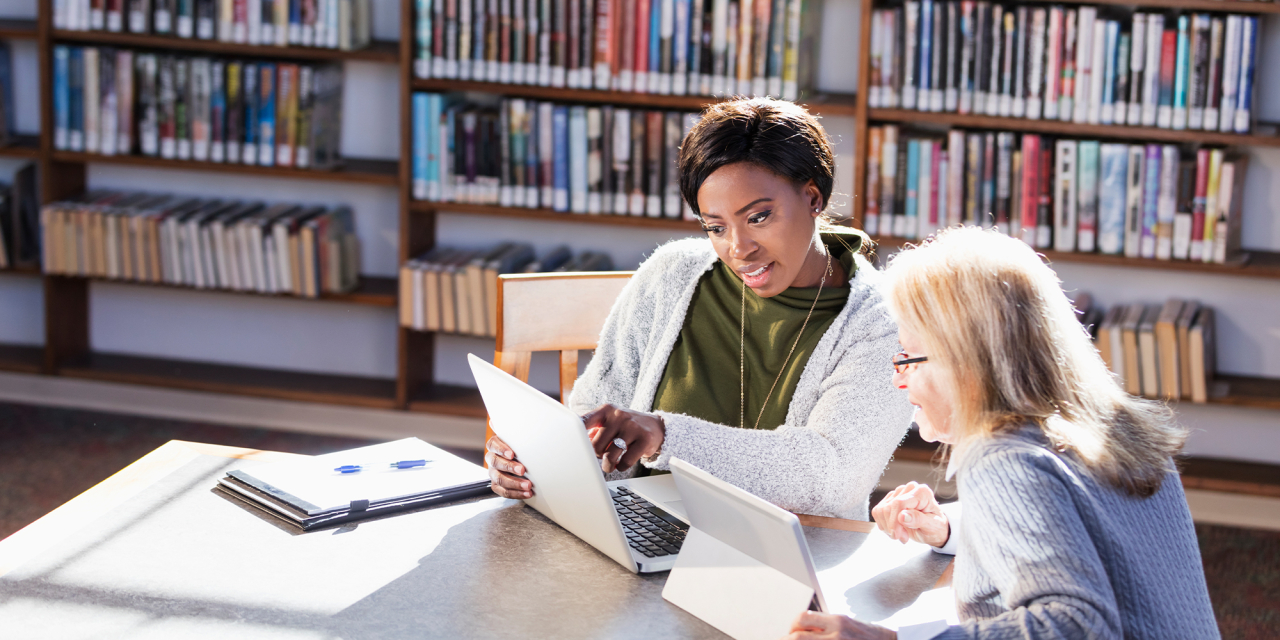 Two people looking at a laptop with rows of books behind them.