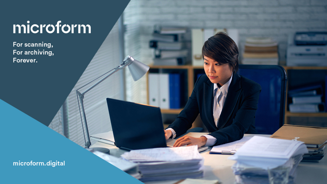 Microform Branding - Woman sat at desk with laptop and stacks of papers.